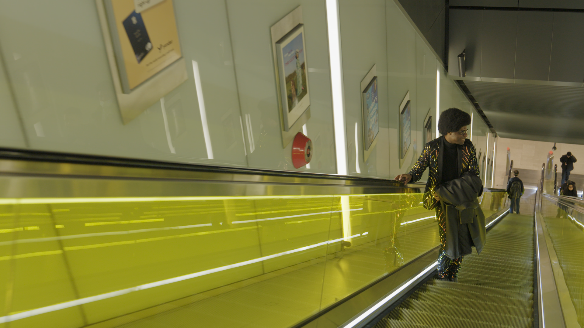 Joel, wearing a glittering suit, smiling as he rides up an escalator
