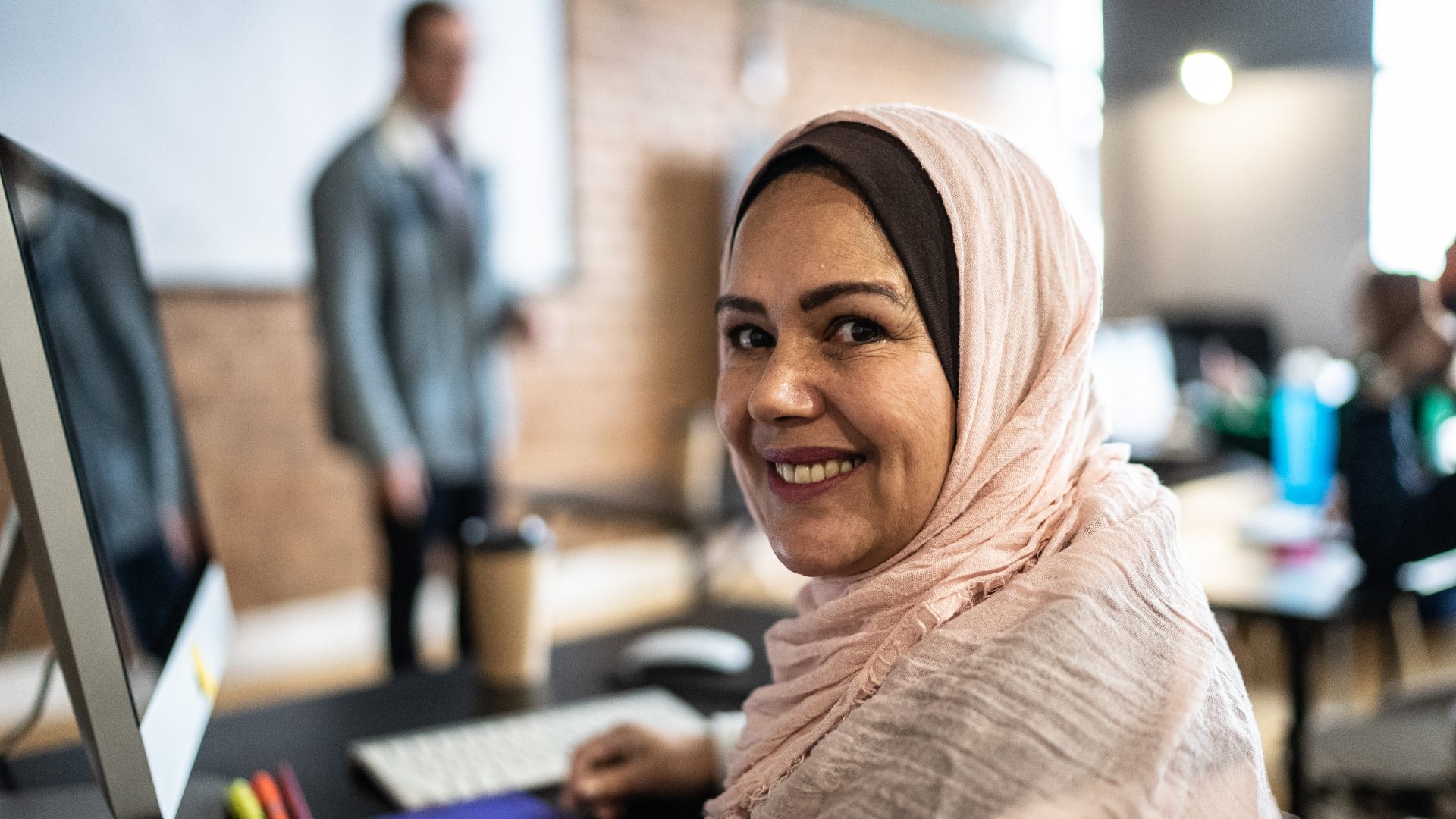 Woman in pink hijab sitting at classroom desk looking over shoulder and smiling